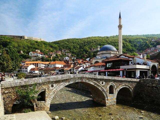 A stone bridge over a creek in a village with a stone tower and red roofs