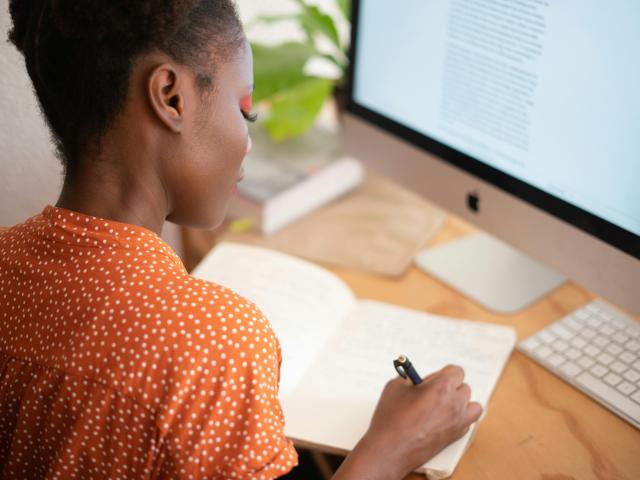 A behind-the-shoulder shot of a woman at her desk writing in a notebook