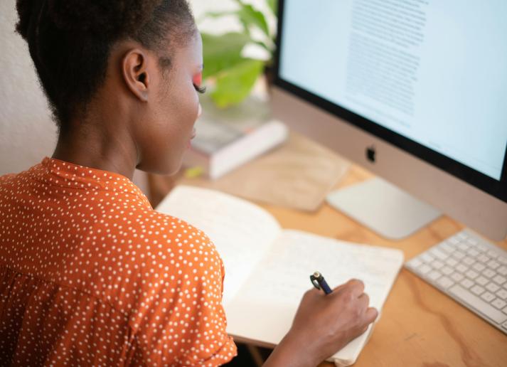 A behind-the-shoulder shot of a woman at her desk writing in a notebook