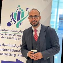 Ahmed Abdul Rahman wearing a suit and standing in front of a sign at a conference smiling at the camera