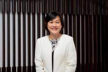 May Tan stands indoors wearing a white jacket, smiling