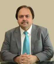 A man sitting with ahnds clasped wearing a suit and a turquoise tie