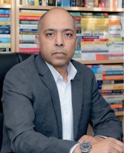 A man wearing a blazer looking at the camera in front of a shelf of books